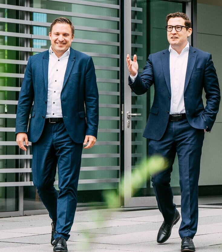 Zwei Männer in Anzügen gehen vor einem modernen Gebäude mit Glas- und Metallfassade entlang, lächeln und unterhalten sich. Im Vordergrund sind Grünpflanzen zu sehen.